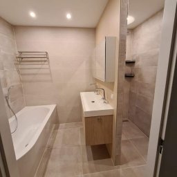 Paweł Gnida - Jasna łazienka po renowacji z wanną, umywalką z szafką i kabiną prysznicową w tle, wykończona płytkami w odcieniach beżu i szarości.