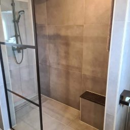 Paweł Gnida - Nowoczesna kabina prysznicowa z czarnymi profilami, szare płytki imitujące beton, wbudowane siedzisko i deszczownica.