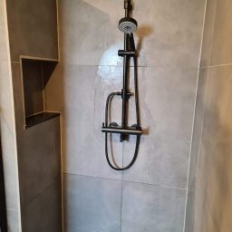 Paweł Gnida - Szara łazienka z kabiną prysznicową i czarną armaturą, wnęka w ścianie wykończona płytkami.