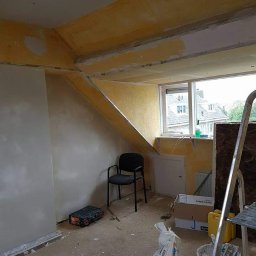 Paweł Gnida - Poddasze w trakcie prac przygotowawczych do malowania: ściany częściowo zagruntowane, widoczne ślady szpachlowania, narzędzia i materiały budowlane porozrzucane na podłodze, krzesło biurowe w rogu...