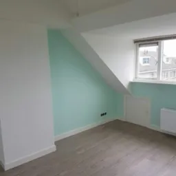 Pomalowane na biało i miętowo poddasze ze skosami, widoczne gniazdka elektryczne, grzejnik i szara podłoga imitująca drewno.
