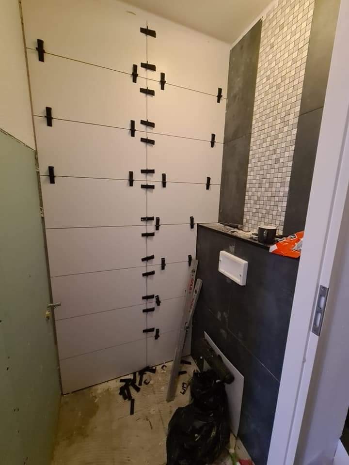 Łazienka w trakcie układania płytek: białe, poziome płytki z czarnymi krzyżykami dystansowymi na ścianie, obok fragment ściany z mozaiką, widoczne narzędzia i poziomica, ślady zaprawy na podłodze.