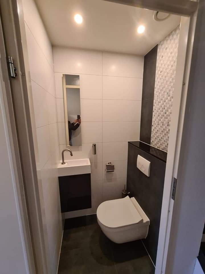 Nowoczesna, mała łazienka z wiszącą toaletą, umywalką nablatową na szafce w kolorze ciemnego brązu, białymi płytkami na ścianach oraz dekoracyjną mozaiką nad zabudową toalety, widok z progu drzwi.