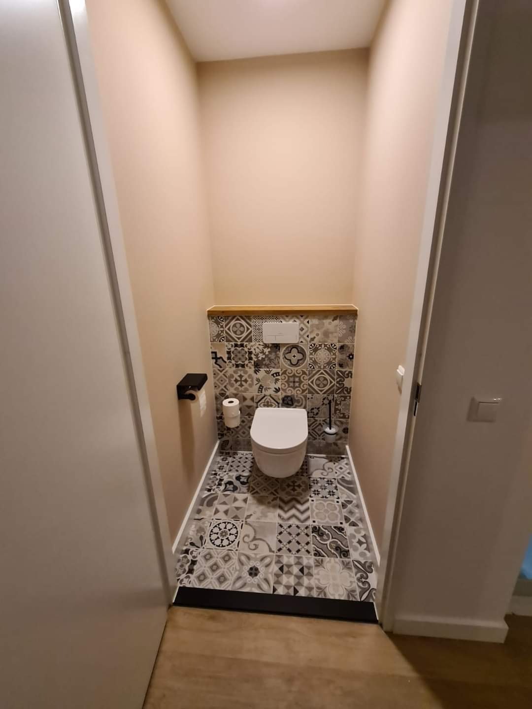 Wąska toaleta z patchworkowymi płytkami w odcieniach szarości i bieli na podłodze i częściowo na ścianie, drewniany blat nad spłuczką, minimalistyczny uchwyt na papier toaletowy.