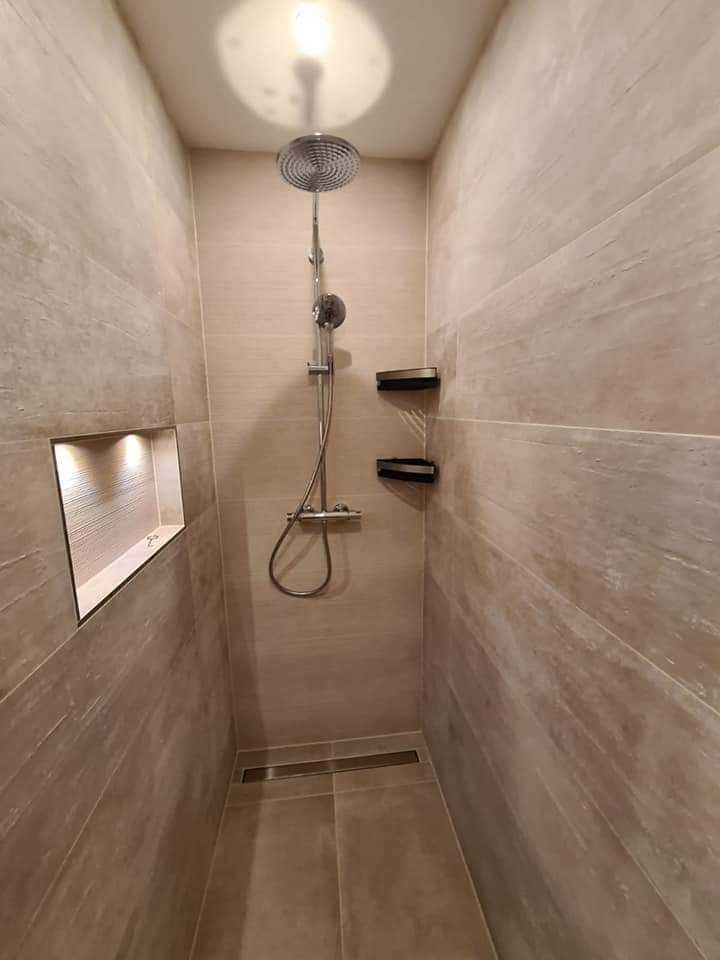 Wąska kabina prysznicowa wyłożona płytkami imitującymi drewno, z deszczownicą i półkami na kosmetyki, oświetlona wnęka w ścianie.