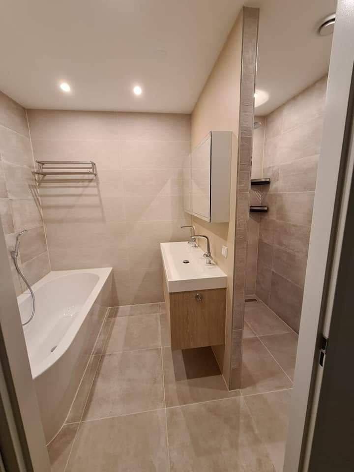 Jasna łazienka po renowacji z wanną, umywalką z szafką i kabiną prysznicową w tle, wykończona płytkami w odcieniach beżu i szarości.