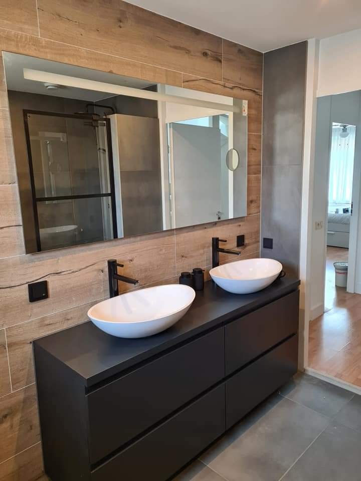 Nowoczesna łazienka z dwoma owalnymi umywalkami nablatowymi, czarną szafką z szufladami, drewnopodobnymi płytkami na ścianie i dużym lustrem z oświetleniem LED.