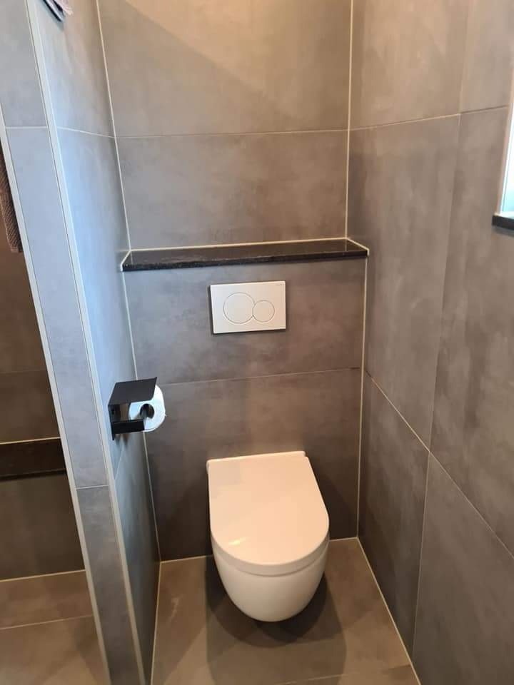 Nowoczesna toaleta podwieszana w łazience wykończonej szarymi płytkami wielkoformatowymi, z czarnym uchwytem na papier toaletowy i półką nad spłuczką.