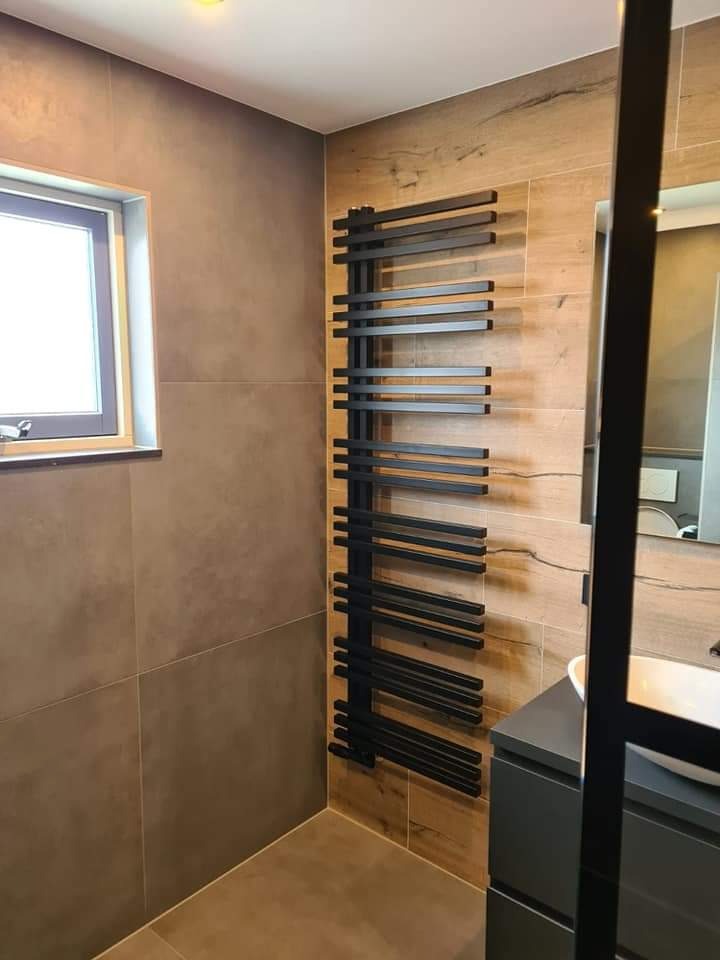 Nowoczesna łazienka z czarnym grzejnikiem drabinkowym na ścianie wyłożonej płytkami imitującymi drewno, obok umywalka nablatowa na szarej szafce, widoczne okno z białymi ramami.