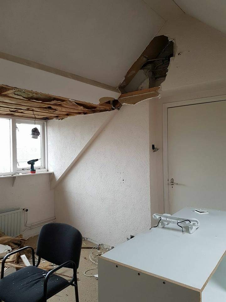 Pokój w trakcie demontażu sufitu, widoczna dziura w suficie z wystającymi elementami konstrukcyjnymi i instalacjami, w tle okno z wkrętarką na parapecie, biurko i krzesło w nieładzie.