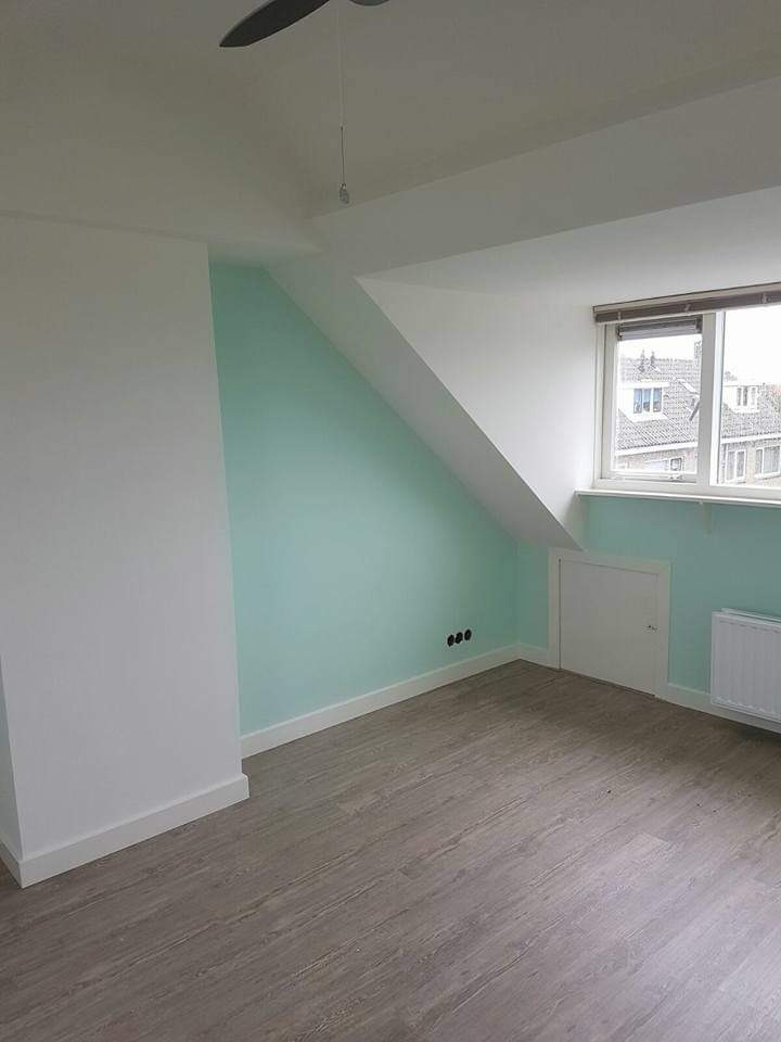 Pomalowane na biało i miętowo poddasze ze skosami, widoczne gniazdka elektryczne, grzejnik i szara podłoga imitująca drewno.