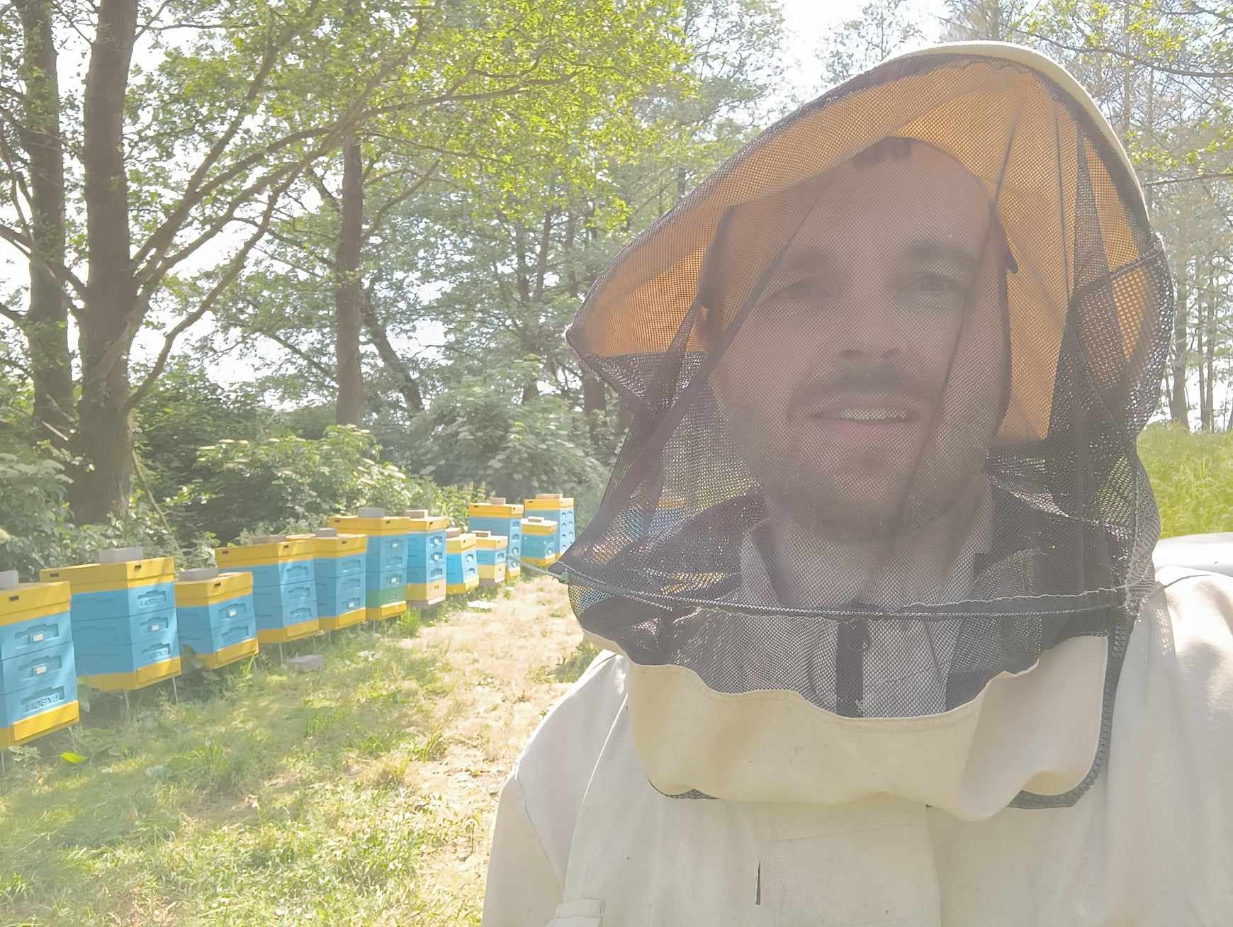 Pszczelarz z Wrocławia w ochronnym stroju i siatce na twarzy, uśmiechnięty, na tle uli ustawionych w rzędzie na łące, widoczne drzewa.