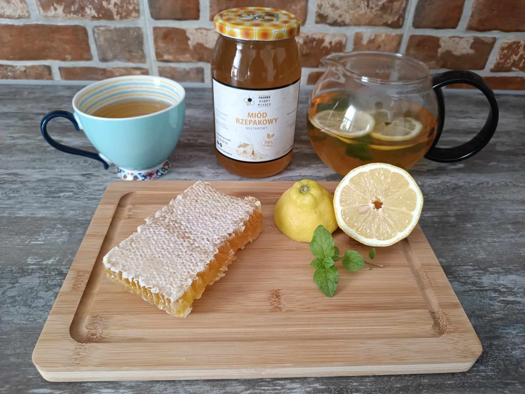 Martwa natura: plaster miodu na desce, słoik miodu rzepakowego, napar z cytryną i miętą w szklanym dzbanku oraz filiżanka herbaty na tle ceglanej ściany.