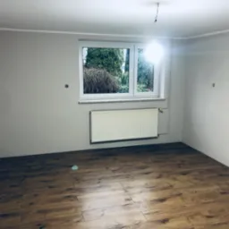 Pomalowane na szaro wnętrze pokoju z ułożonymi panelami podłogowymi, białym grzejnikiem pod oknem i wystającym kablem od lampy.