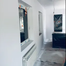 Wnętrze w trakcie odświeżania: świeżo pomalowane białe ściany, zakryte folią meble, zabezpieczona podłoga kartonem oraz grzejnik pod oknem.