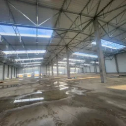 Wnętrze hali magazynowej w trakcie budowy: betonowa posadzka z kałużami, stalowa konstrukcja dachu z świetlikami, betonowe słupy. Widok z poziomu posadzki.