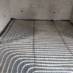 Białe rurki ogrzewania podłogowego ułożone na czarnej macie w surowym pomieszczeniu z białych bloczków. Widoczne pionowe rurki instalacji.