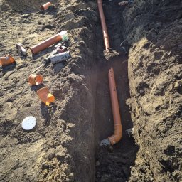 FHU INSTABUD Sebastian Olczak - Wykonanie rowu pod instalację kanalizacyjną z pomarańczowymi rurami PCV. Widoczne narzędzia i elementy instalacji w wykopie ziemnym.