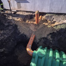 FHU INSTABUD Sebastian Olczak - Instalacja kanalizacyjna zewnętrzna: zielony zbiornik i pomarańczowe rury w wykopie, widoczna łopata i fragment ściany budynku. Prace ziemne w toku.