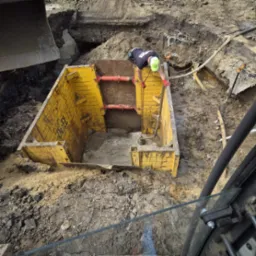 Pracownik w żółtej klatce zabezpieczającej wykop, koparka w tle. Praca ziemne przy fundamentach z użyciem szalunków. Wykop zabezpieczony przed osunięciem.