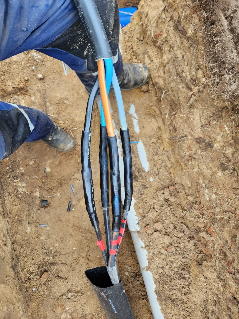 Podłączenie kabla elektrycznego w wykopie ziemnym. Widoczne kolorowe przewody i elementy izolacyjne. Detal pracy instalatora przyłącza elektrycznego.