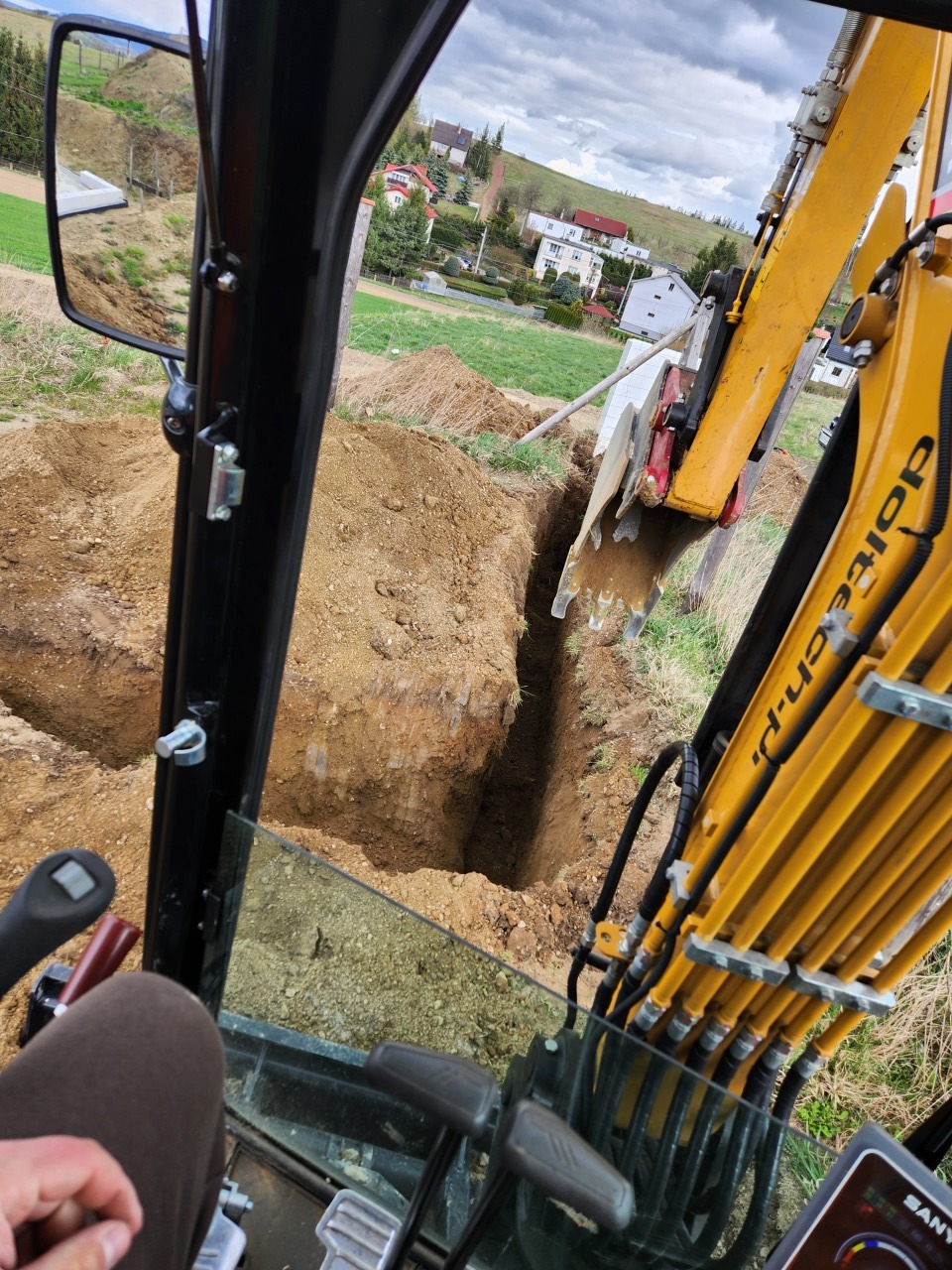 Kopanie rowu koparką SANY na tle zabudowy jednorodzinnej. Widok z kabiny operatora, na pierwszym planie dłoń. Praca w toku, świeżo wykopany rów.