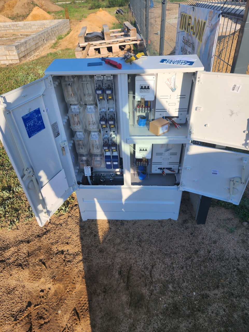 Otwarta skrzynka elektryczna z bezpiecznikami i licznikami na zewnątrz, widoczne narzędzia i elementy instalacji. W tle teren budowy i ogrodzenie.