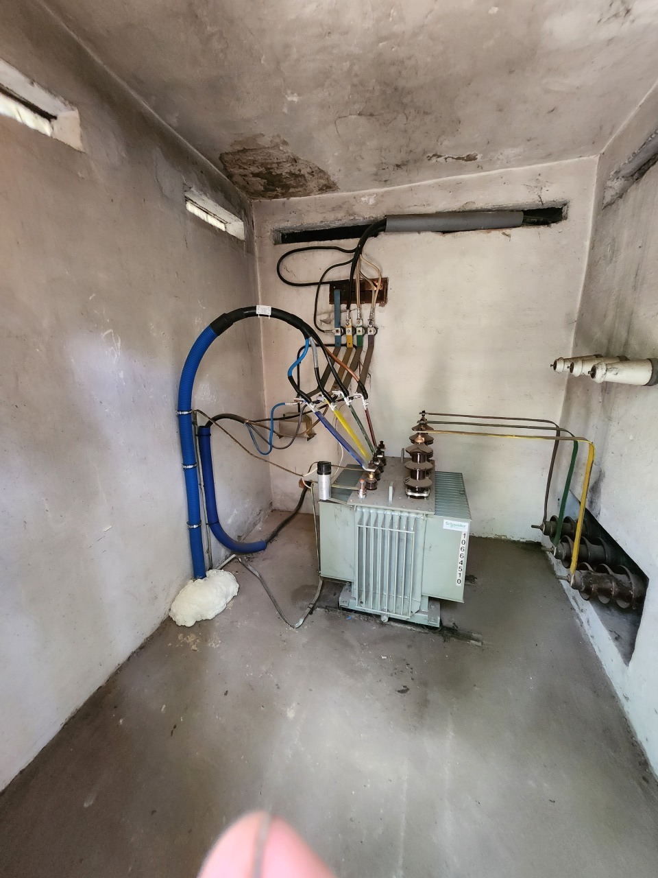 Wnętrze pomieszczenia z zainstalowanym transformatorem elektrycznym, widoczne przewody i kable. Surowe ściany i podłoga. Instalacja w małym pomieszczeniu.