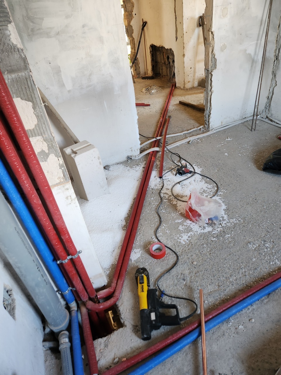 Instalacja rur grzewczych (czerwone i niebieskie) w surowym wnętrzu. Widoczne narzędzia, fragment podłogi i ściany w trakcie prac instalacyjnych.