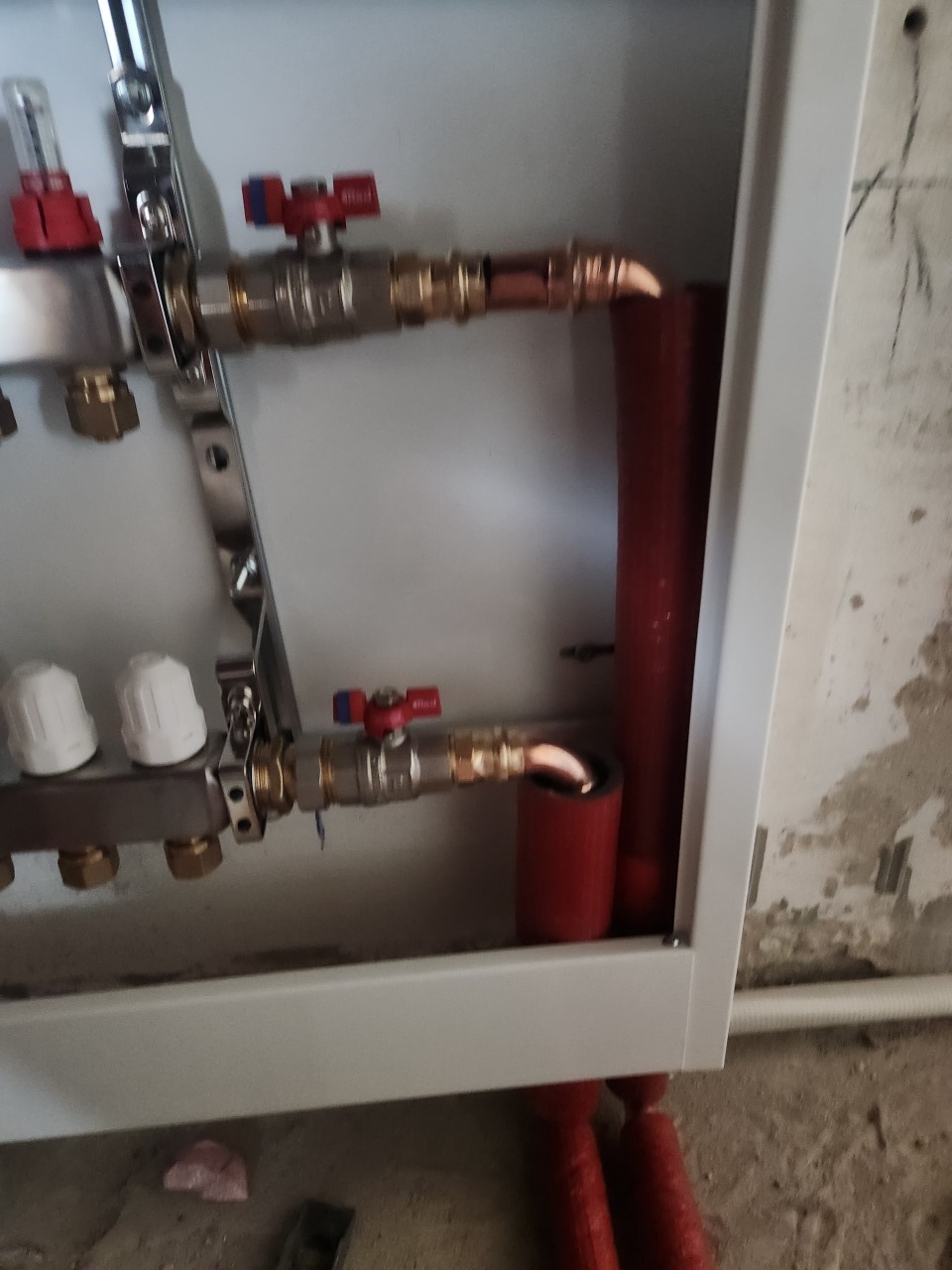 Rozdzielacz ogrzewania podłogowego w szafce, z widocznymi zaworami, rurami miedzianymi i czerwonymi rurami grzewczymi. Szare tło ściany w tle.