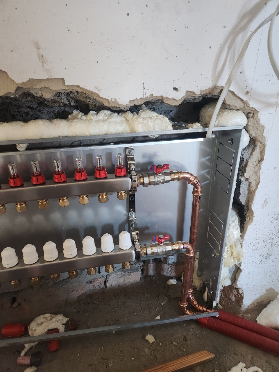 Rozdzielacz ogrzewania podłogowego z miedzianymi rurami i elementami regulacyjnymi, zamontowany na ścianie z izolacją piankową. Widoczne czerwone rury.