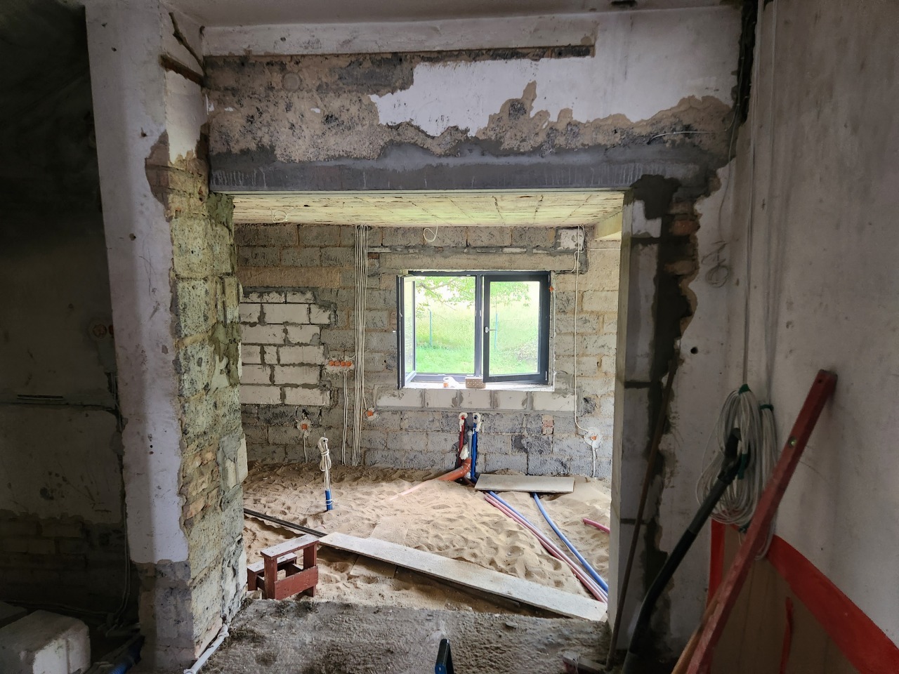 Wnętrze w trakcie remontu: murowane ściany z widoczną instalacją elektryczną i hydrauliczną, piasek na podłodze, okno z widokiem na zieleń. Prace murarskie i instalacyjne w toku.
