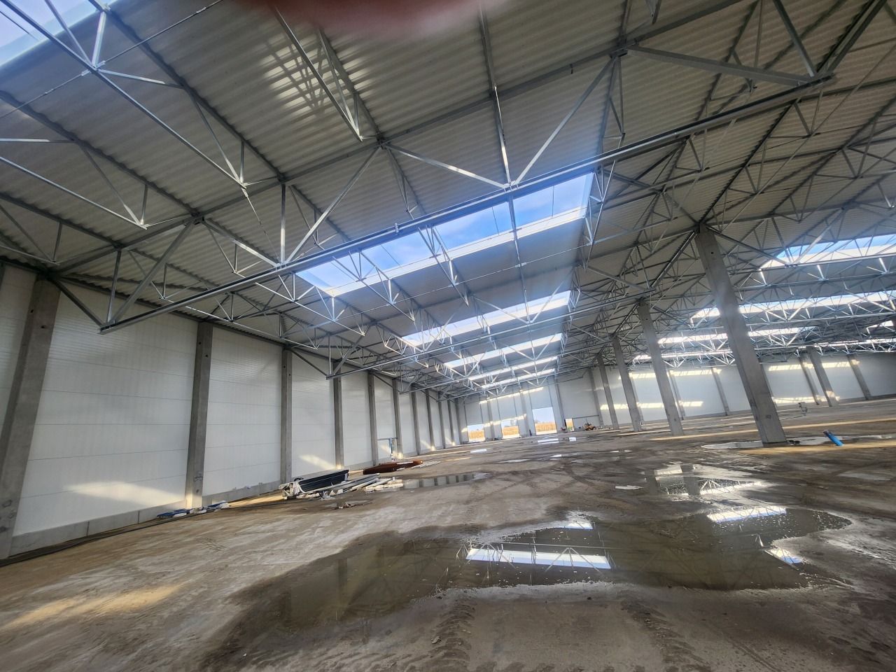 Wnętrze hali magazynowej w trakcie budowy. Widoczne stalowe elementy konstrukcyjne dachu i betonowa posadzka z refleksami światła. Hala oświetlona naturalnym światłem.