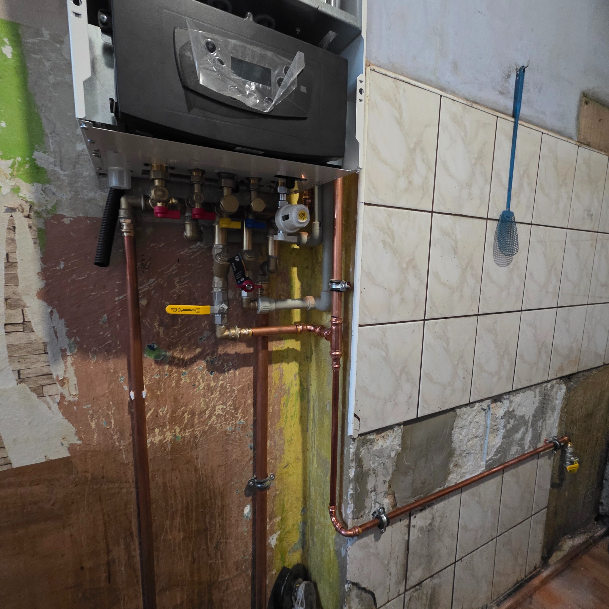 Nowy piec gazowy zamontowany na ścianie z częściowo usuniętymi płytkami, widoczne miedziane rury instalacji grzewczej z zaworami. W tle łapka na muchy.