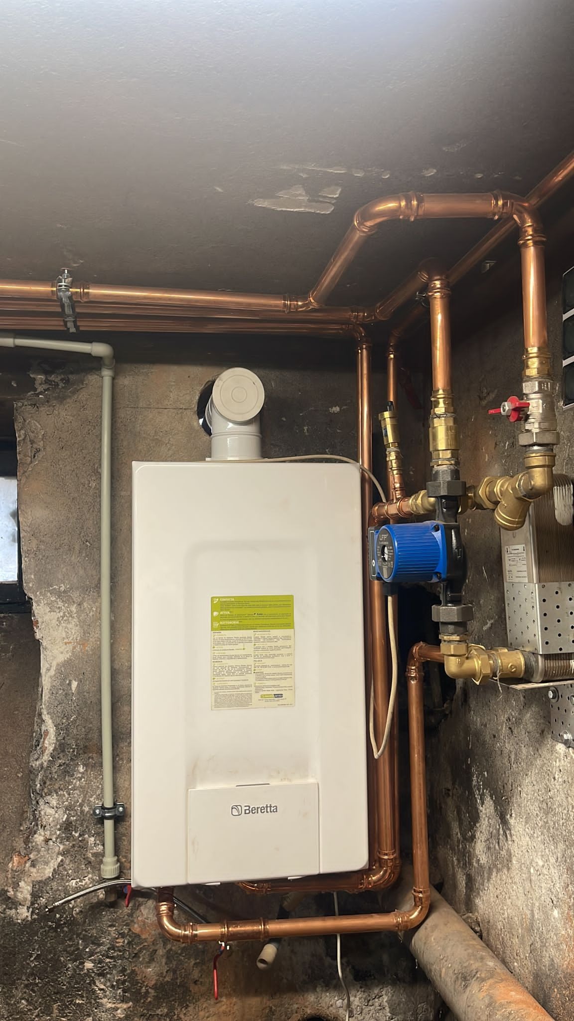 Nowoczesny piec gazowy Beretta z miedzianą instalacją, zamontowany na tle surowej ściany. Widoczne złącza i zawory, tworzące skomplikowany system.