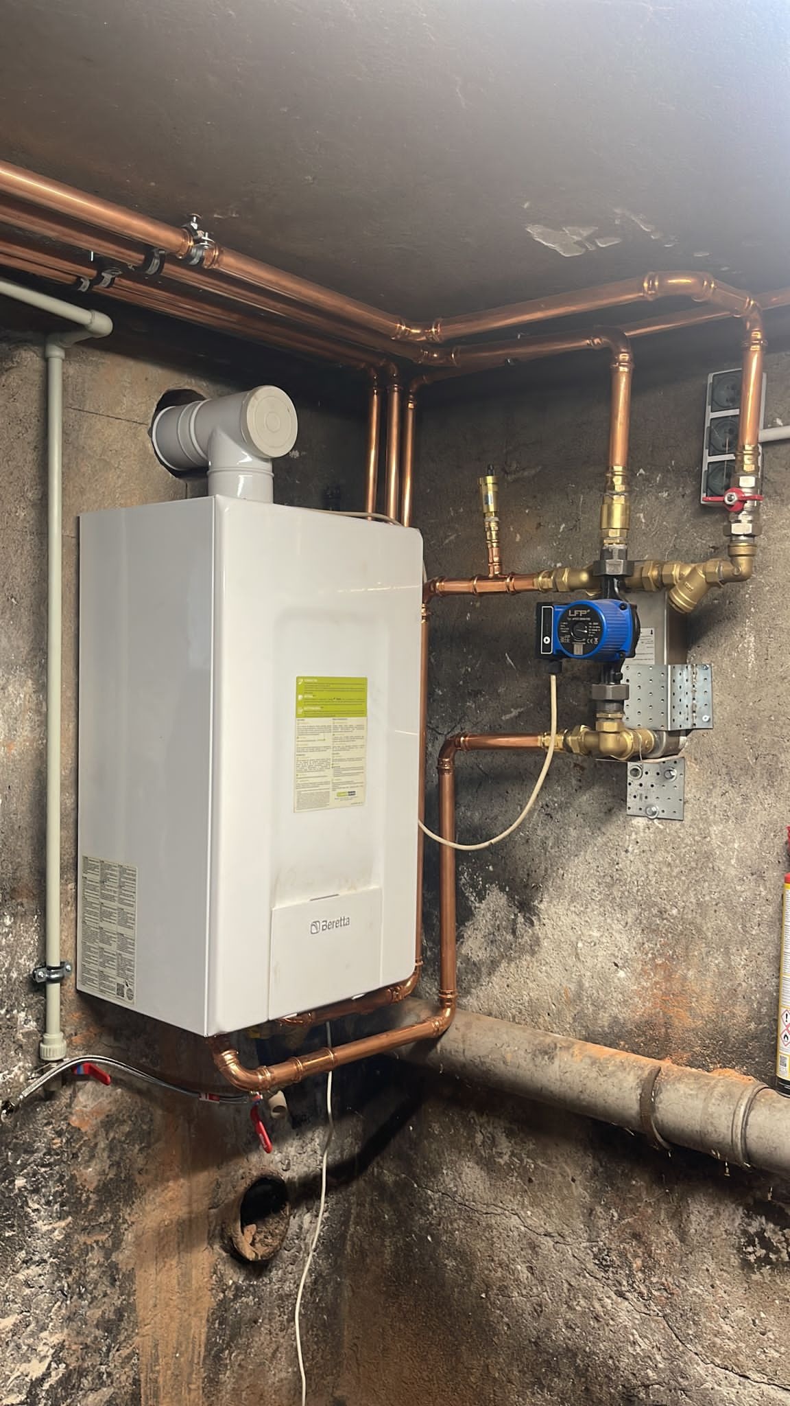 Biały piec gazowy Beretta z miedzianą instalacją grzewczą na tle surowej, betonowej ściany. Widoczne rury i zawory.