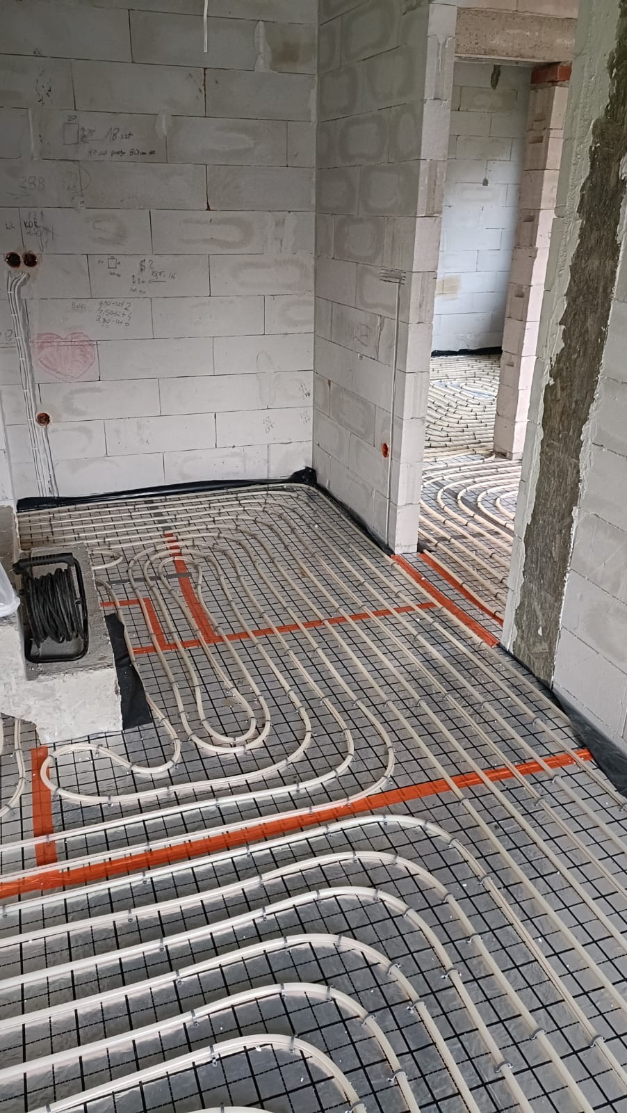 Instalacja ogrzewania podłogowego z rurami w kolorze kremowym, zamocowanymi do czarnej siatki na folii. Widoczne ściany z białych bloczków. Pomieszczenie w trakcie budowy.