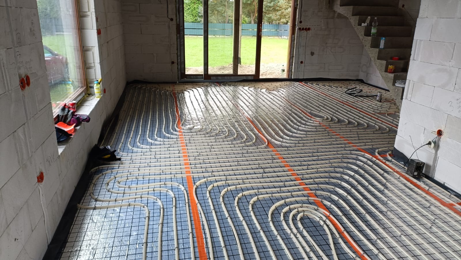 Białe rury ogrzewania podłogowego na czarnej macie, ułożone w pętle w surowym wnętrzu domu z oknem i schodami. Pomarańczowe akcenty rur.