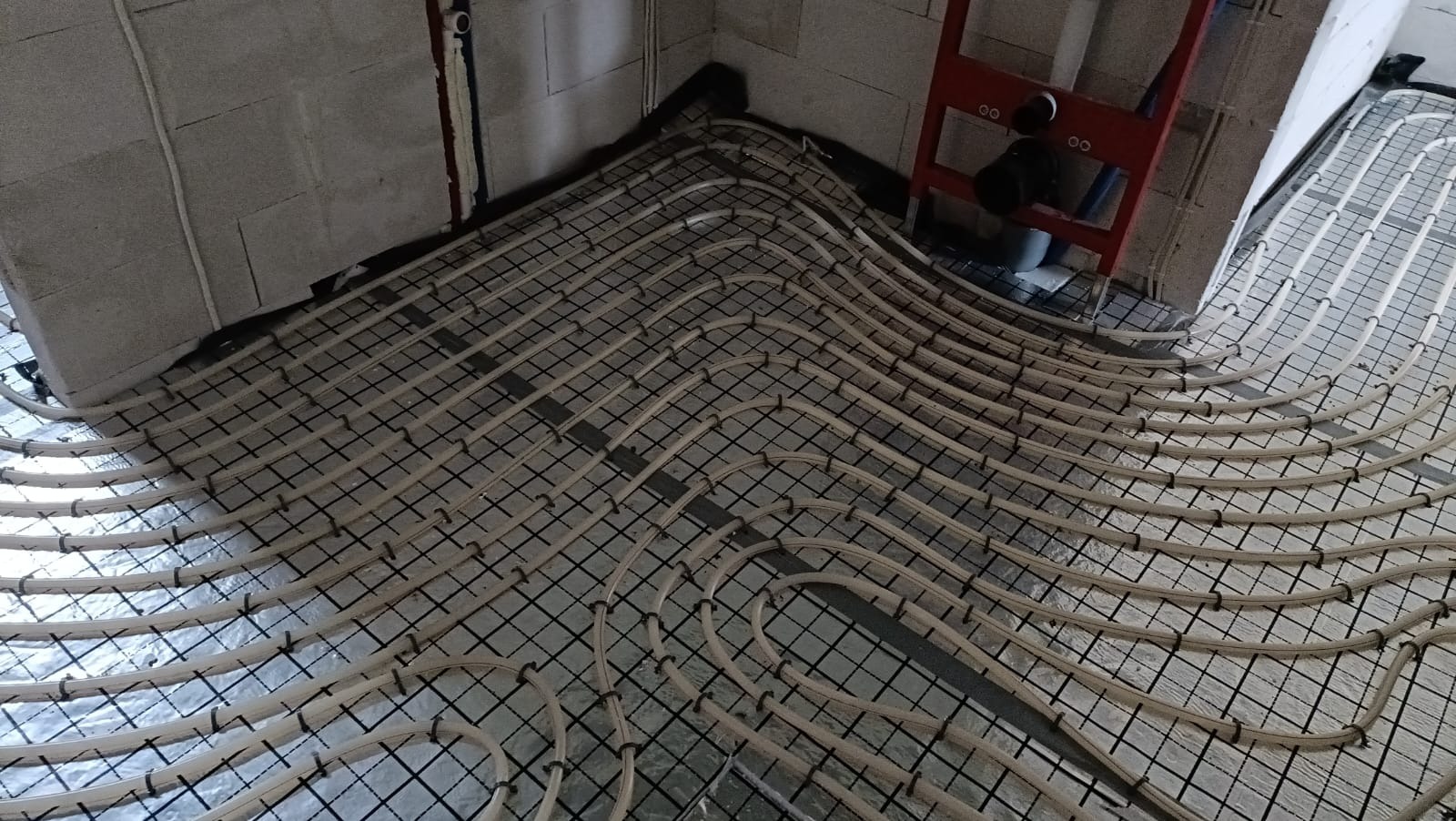 Instalacja ogrzewania podłogowego w surowym wnętrzu. Rury ułożone na siatce, widoczne ściany z bloczków i elementy instalacji sanitarnej.