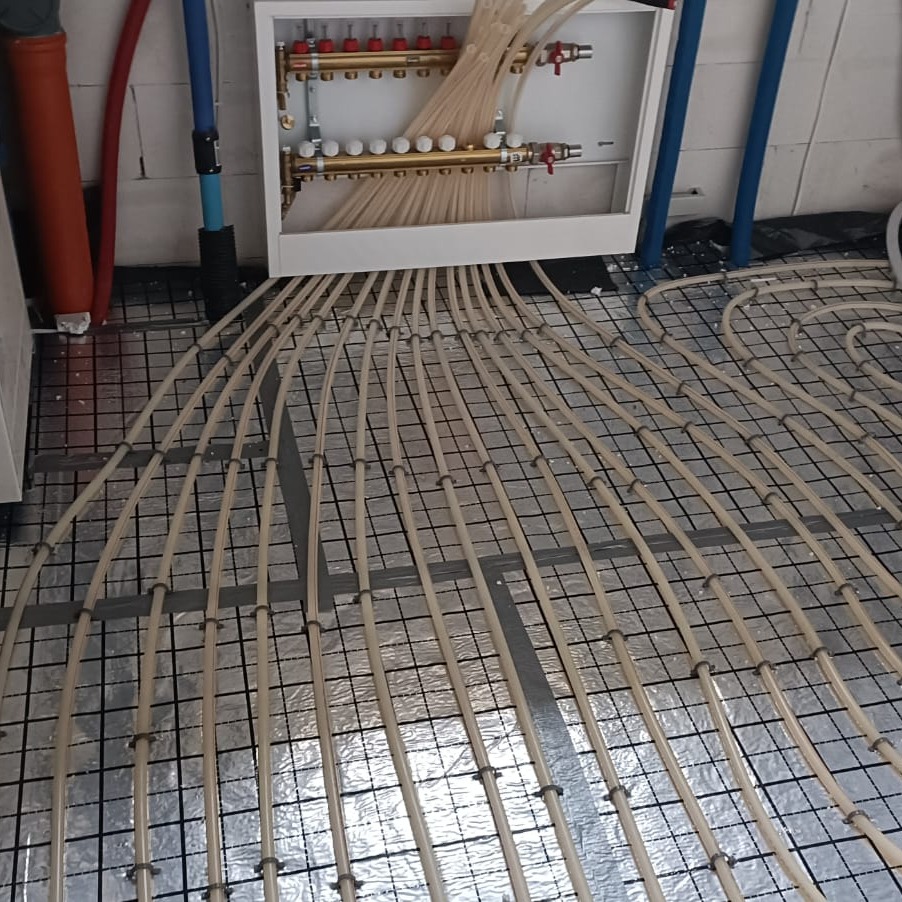 Montaż ogrzewania podłogowego: rury rozprowadzone na folii izolacyjnej z zamocowaniami, widoczny rozdzielacz w białej obudowie. Układ spiralny rur.