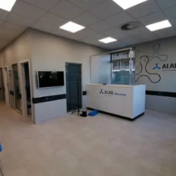 Jasne, przestronne wnętrze laboratorium ALAB z recepcją, niebieskimi krzesłami dla pacjentów i telewizorem na ścianie. Na ścianie logo firmy i dystrybutor wody.