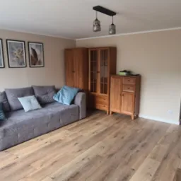 Jasne panele podłogowe w salonie z szarą sofą i drewnianymi meblami, widok na osobę sprzątającą w kuchni w tle.