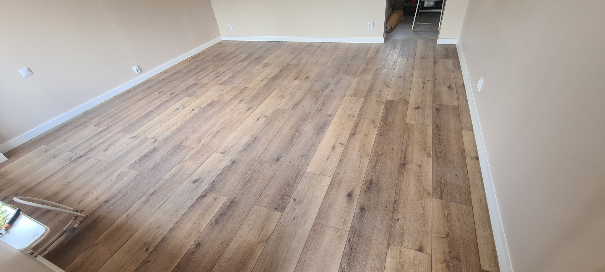 Jasne panele podłogowe ułożone w pustym pomieszczeniu z kremowymi ścianami i białymi listwami przypodłogowymi, widoczne narzędzia pracy w rogu.