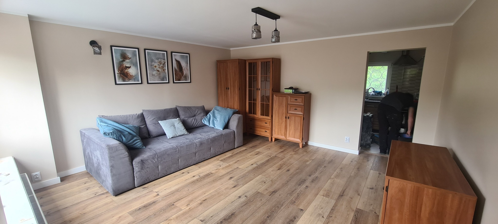 Jasne panele podłogowe w salonie z szarą sofą i drewnianymi meblami, widok na osobę sprzątającą w kuchni w tle.