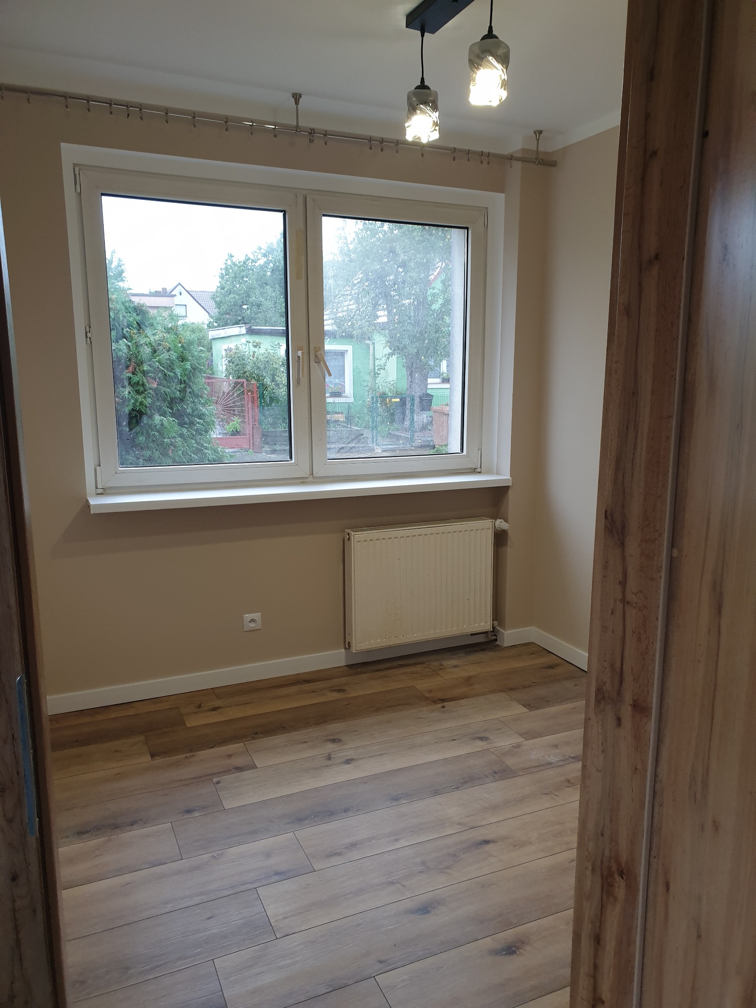 Jasne wnętrze pokoju z nową podłogą z paneli imitujących drewno, widok zza otwartych drzwi, okno z widokiem na ogród, grzejnik pod oknem.