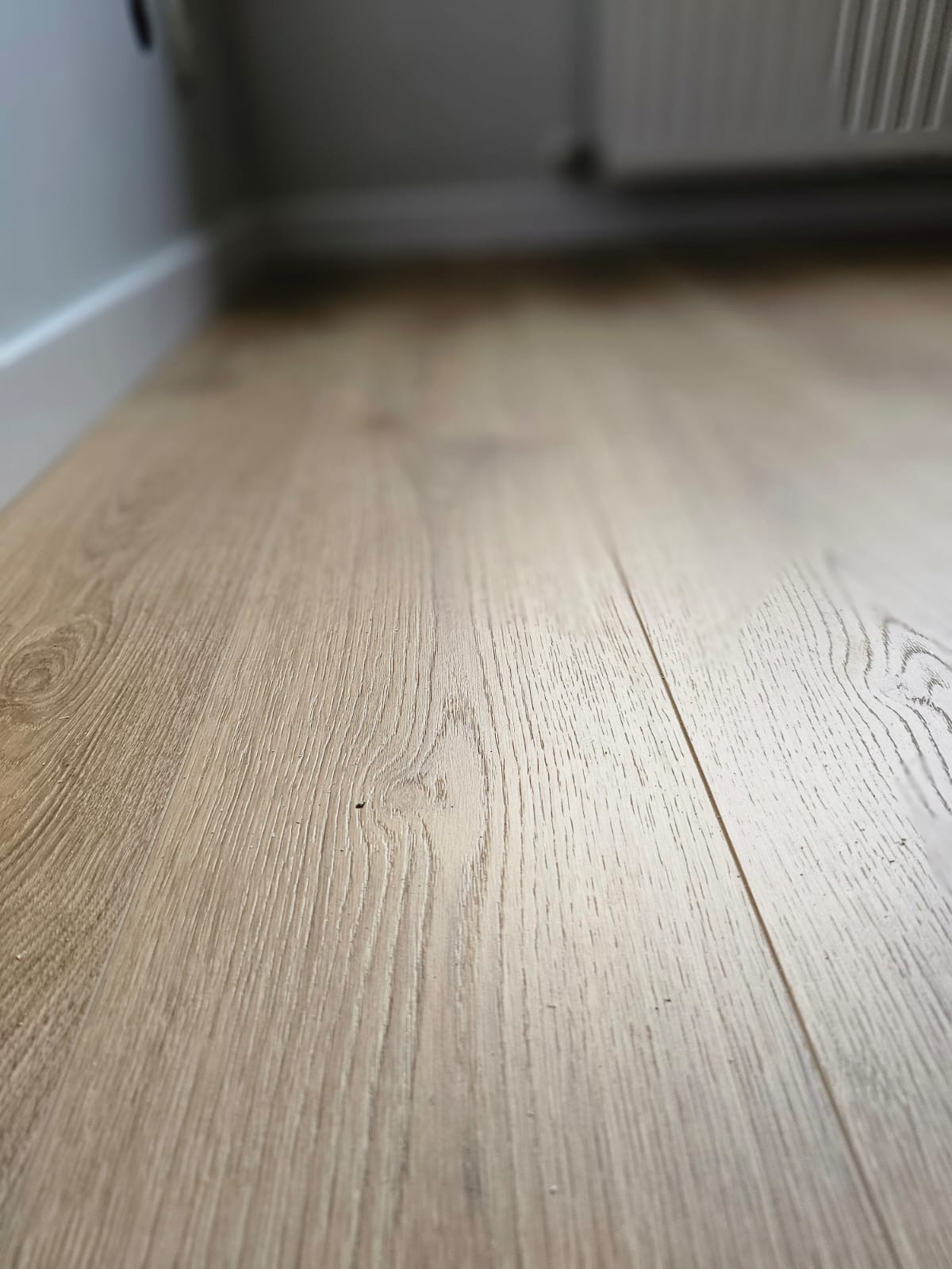 Jasne panele podłogowe imitujące drewno, ułożone wzdłuż ściany z białą listwą przypodłogową i grzejnikiem w tle.