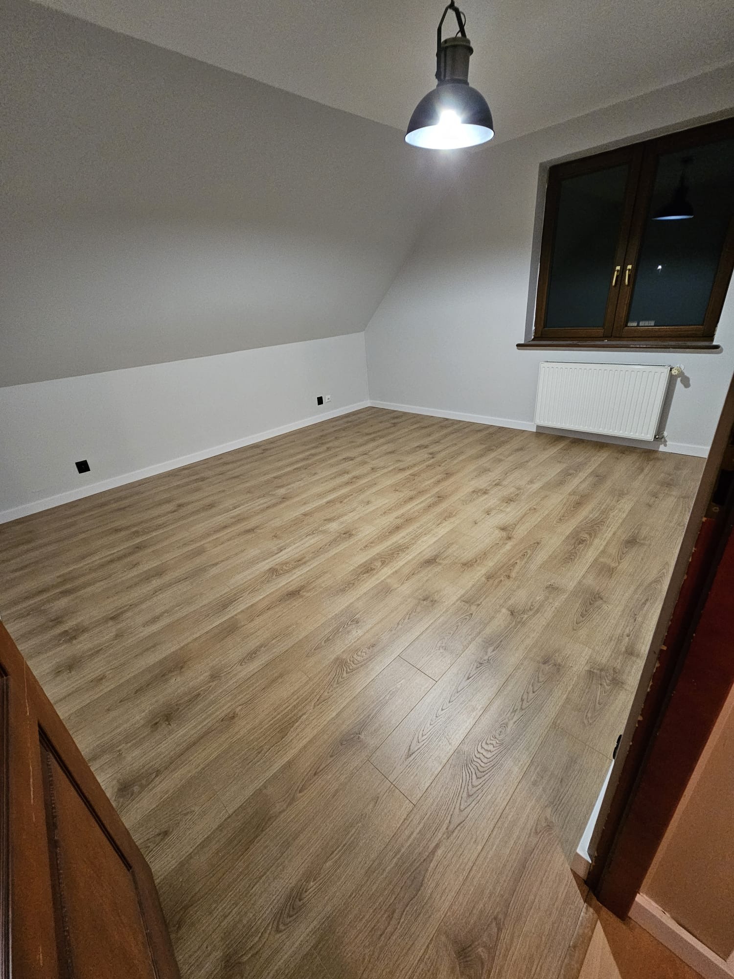 Jasne panele podłogowe w pokoju na poddaszu z oknem i grzejnikiem, widok zza otwartych drzwi, oświetlone wiszącą lampą w stylu industrialnym.