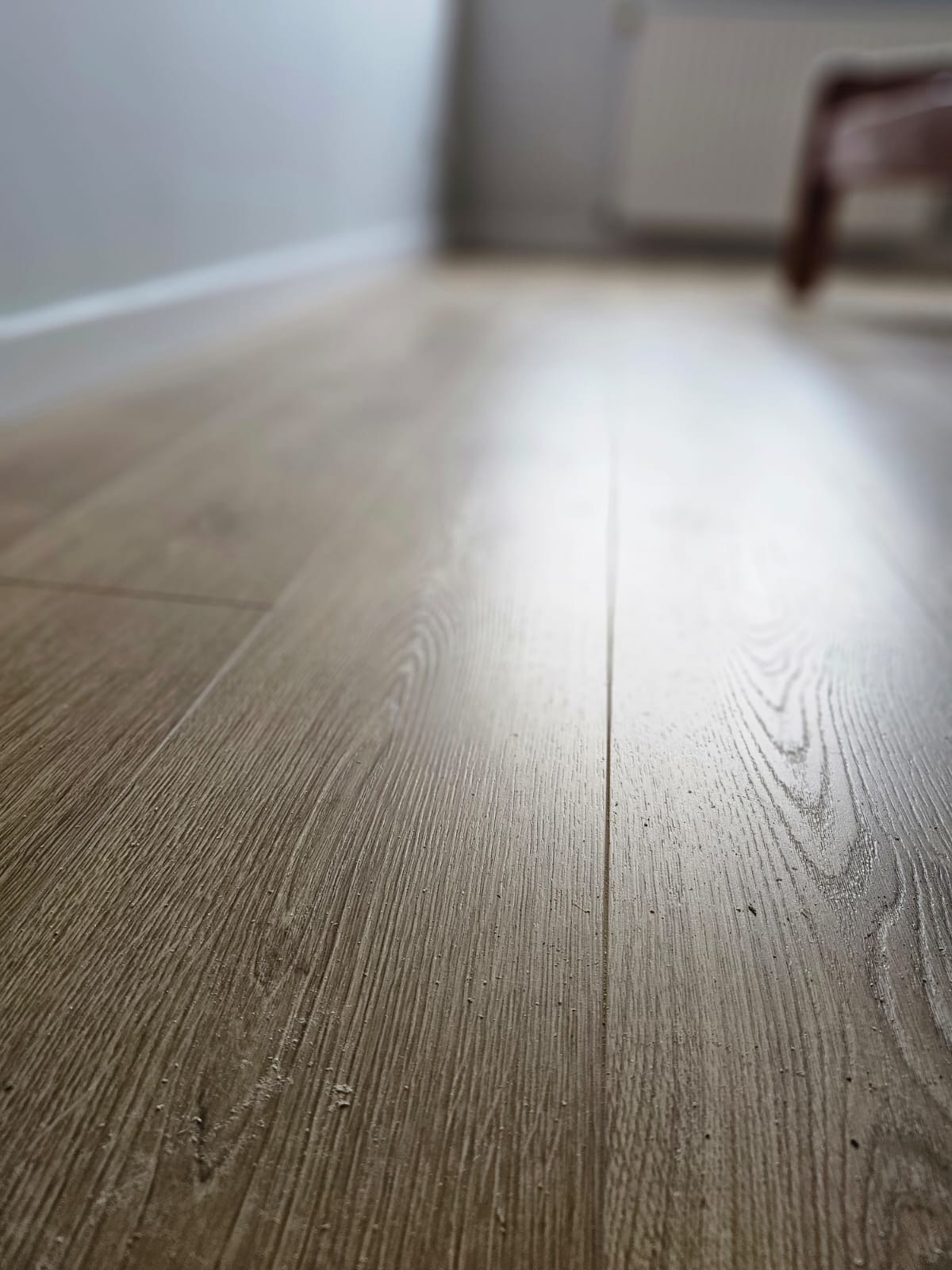Jasnobrązowa podłoga z paneli laminowanych, ułożona wzdłuż, z widoczną strukturą drewna i delikatnym połyskiem, z fragmentem białej listwy przypodłogowej i rozmazanym tłem z meblem w tle.