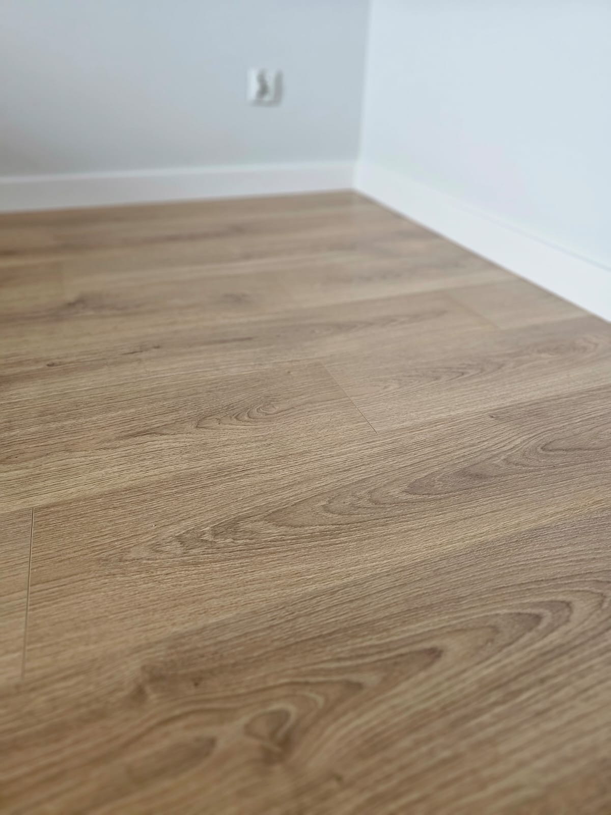 Jasnobrązowe panele podłogowe ułożone w rogu pomieszczenia z białymi listwami przypodłogowymi i gniazdkiem elektrycznym na ścianie.