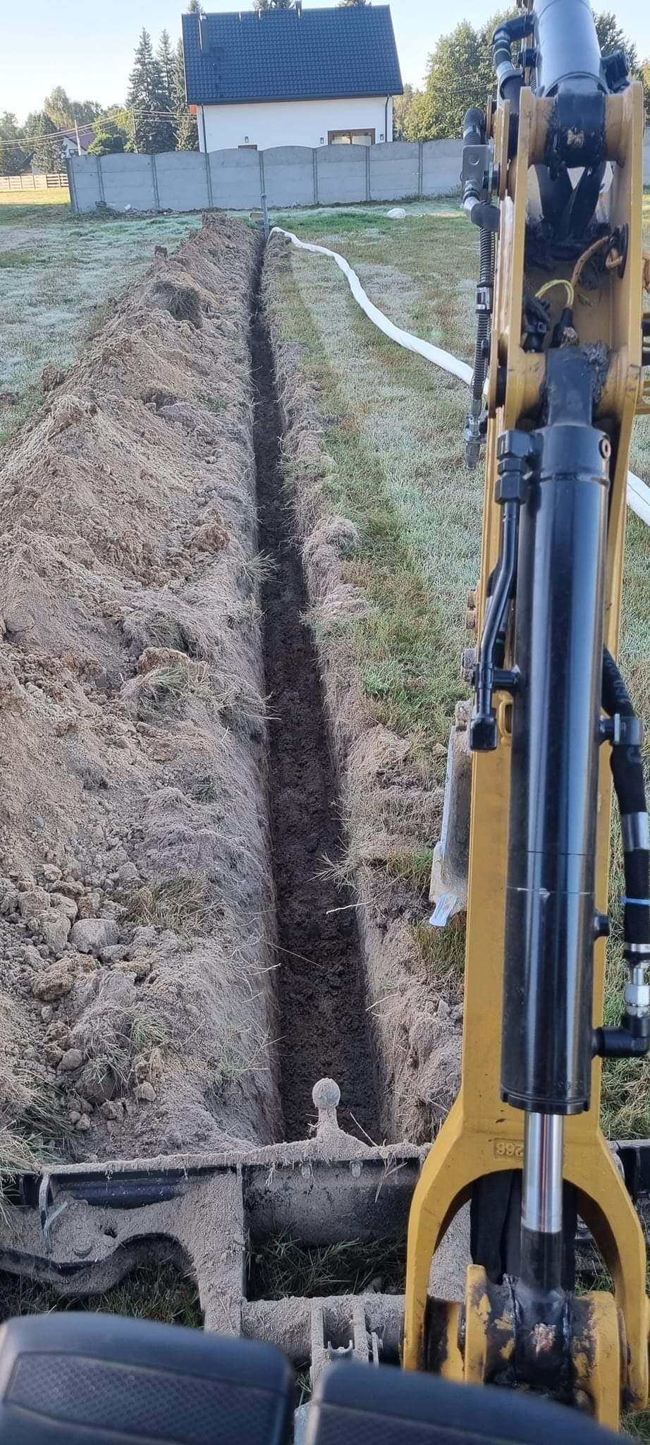 Wąski wykop w ziemi, wykonany koparką, widoczny element maszyny i biała rura ułożona wzdłuż wykopu na trawie pokrytej szronem, w tle dom jednorodzinny i drzewa.